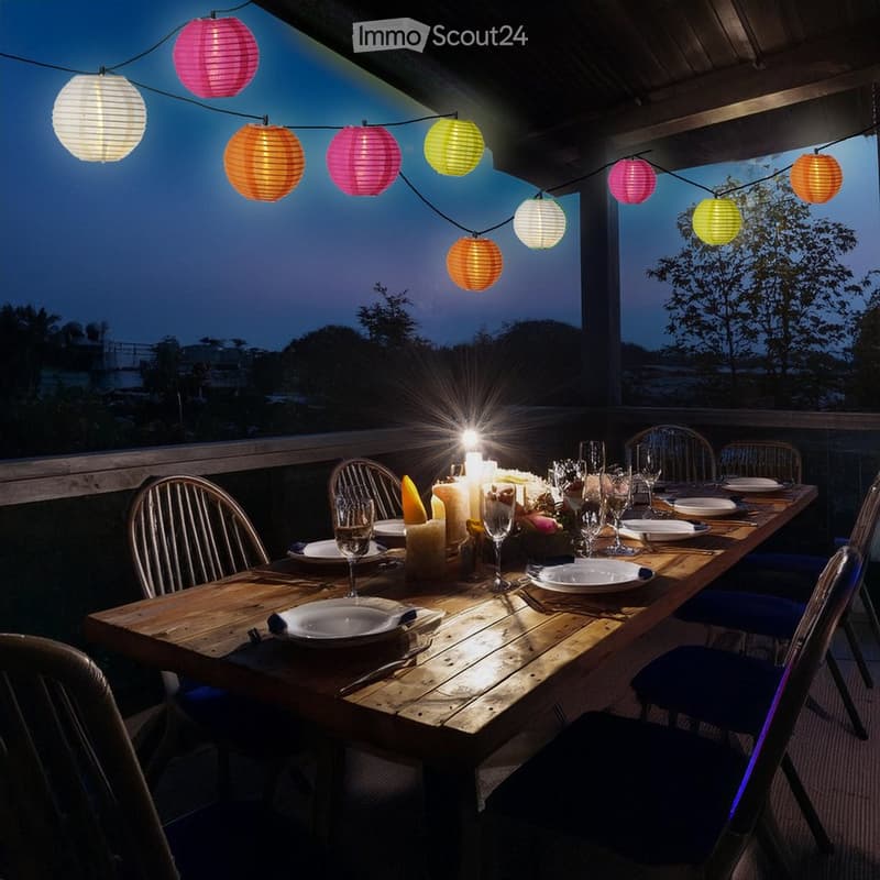 A wooden dining table set for a meal with plates, glasses, and candles, surrounded by colorful paper lanterns and blue chairs, set against a twilight backdrop with trees.