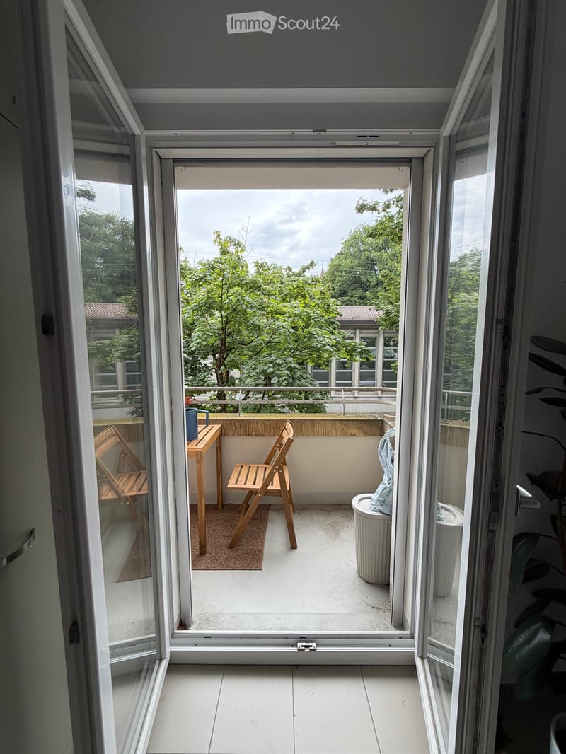 Balcony with wooden table, two chairs, a cup, and a trash can, overlooking a green area with trees.