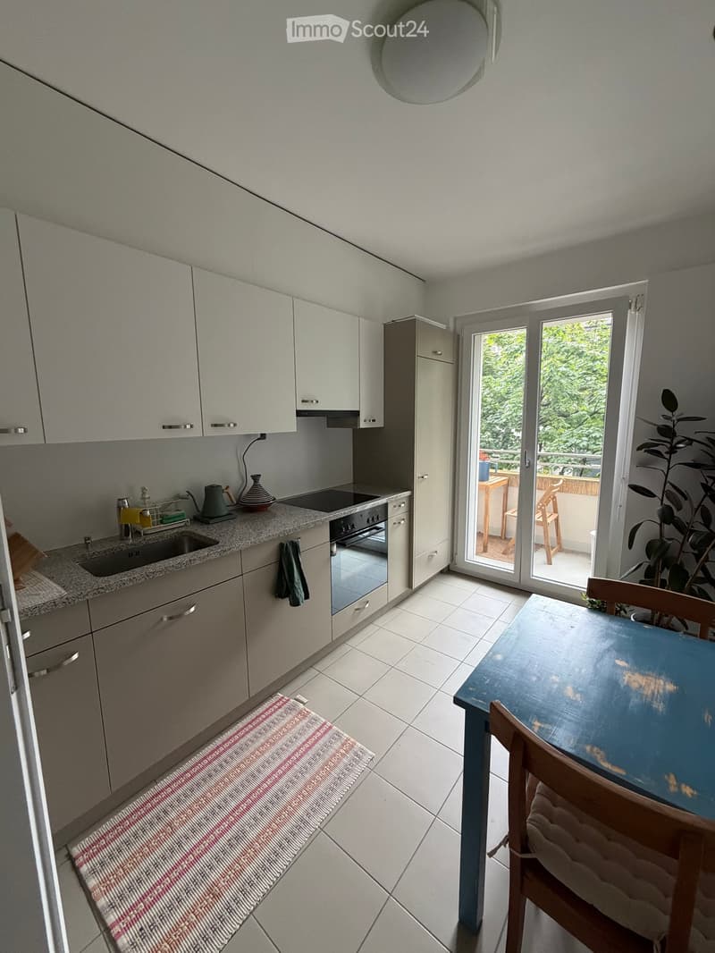 Modern kitchen with white cabinets, granite countertop, built-in oven, sink, and a small dining area with a table and chair. A sliding glass door leads to a balcony.