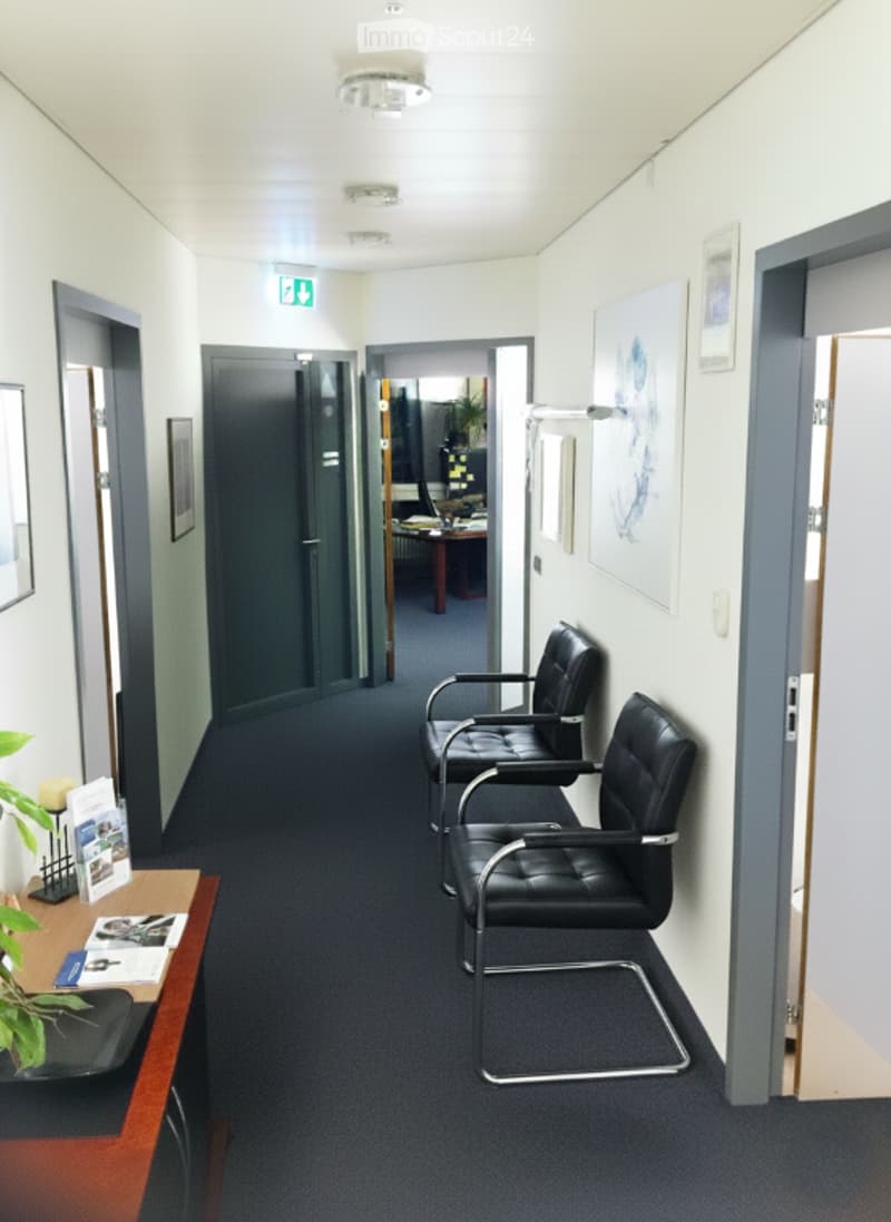 A clean, modern hallway with a dark blue carpet, white walls, and several doors. Two black leather chairs are positioned against the wall, and a small wooden table with brochures and a plant is visible on the left side.