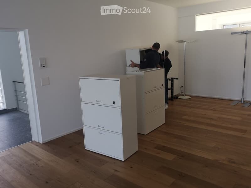 Empty room with wooden flooring, white walls, and large windows. Two filing cabinets are present, but the room is otherwise unfurnished.