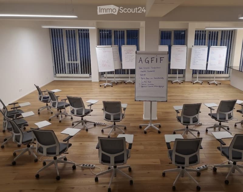 Modern office meeting room with wooden flooring, large windows with blinds, multiple ergonomic chairs, small tables, and flipcharts.