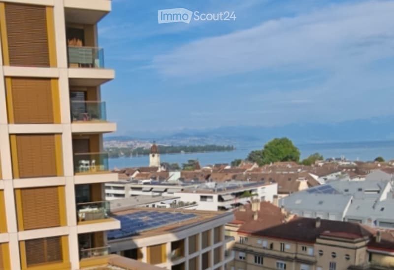 View from a balcony showing a cityscape with rooftops, a lake, and distant mountains.