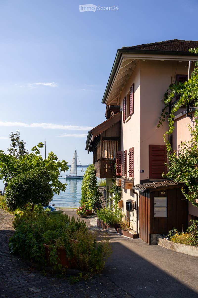 Two-story residential building with a balcony overlooking a lake, surrounded by greenery and a sailboat in the background.