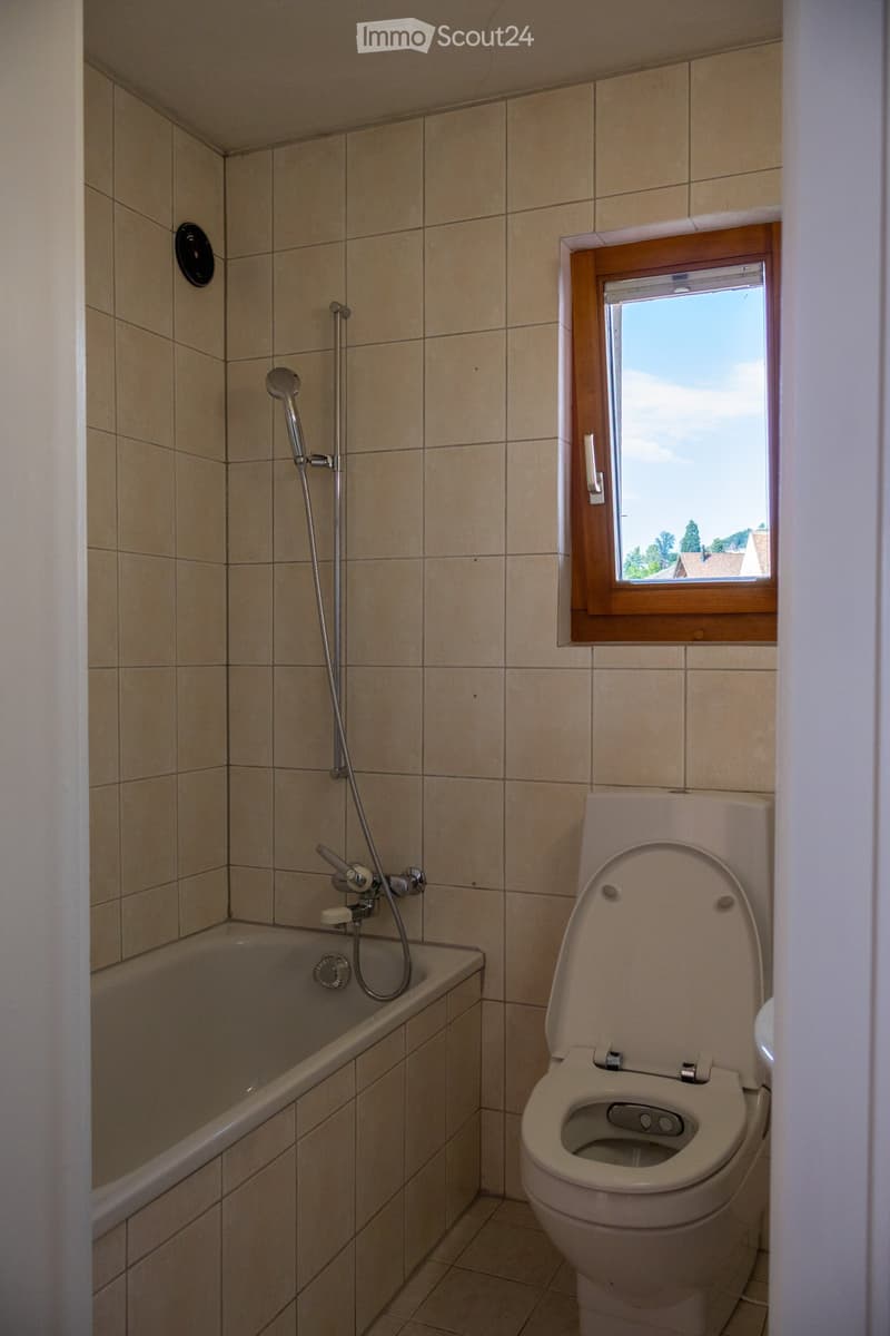 Bathroom with beige tiled walls and floor, featuring a bathtub with a handheld showerhead, a toilet, and a window with wooden frame offering a view of the outdoors.