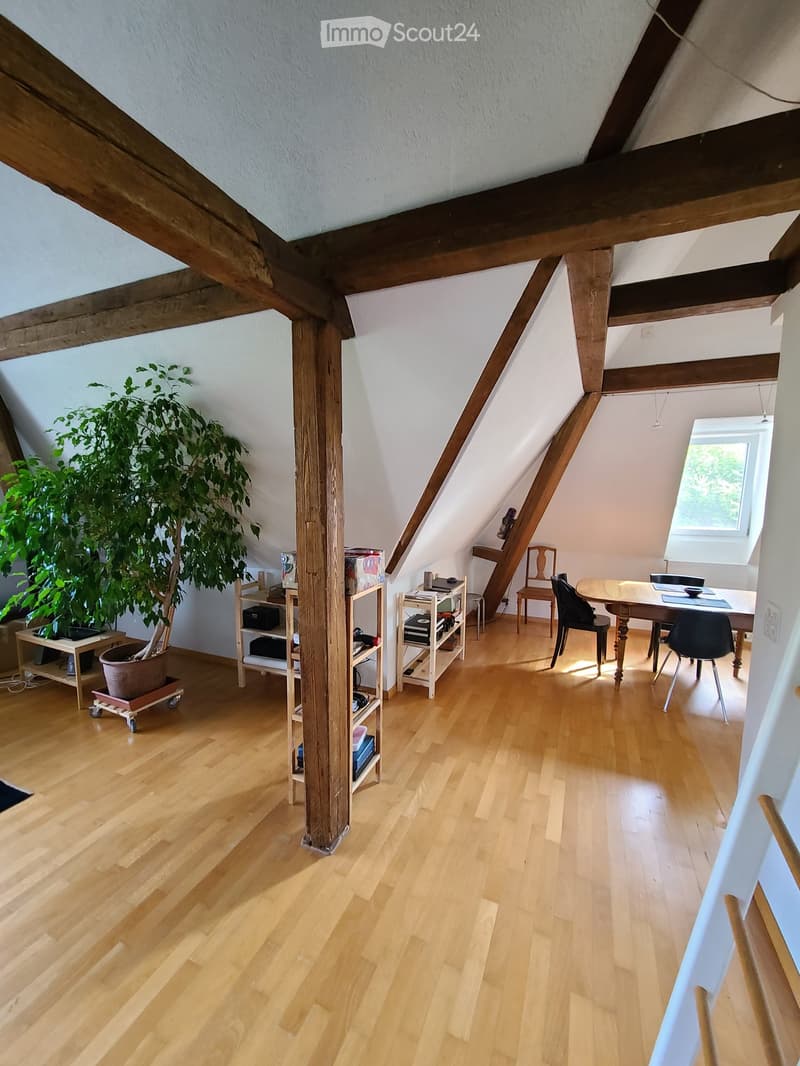 Bright attic room with wooden floors, slanted wooden ceiling beams, large window, and modern furniture including a dining table, chairs, shelves, and a potted plant.