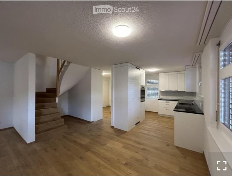 Open-plan space with a staircase leading to an upper level, a kitchen area with white cabinets and a black countertop, and wooden flooring throughout.