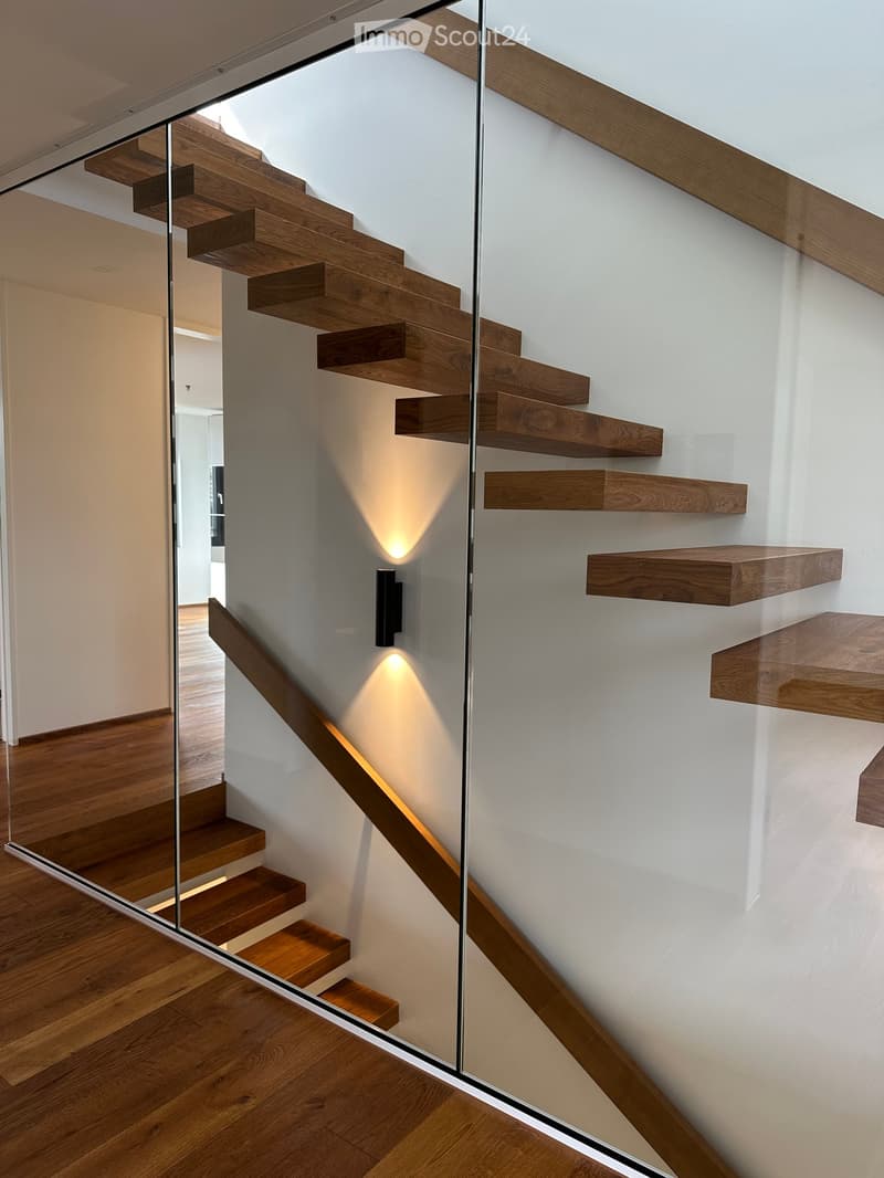 Modern staircase with wooden steps and a glass railing, illuminated by wall-mounted spotlights, leading to an open area with wooden flooring.