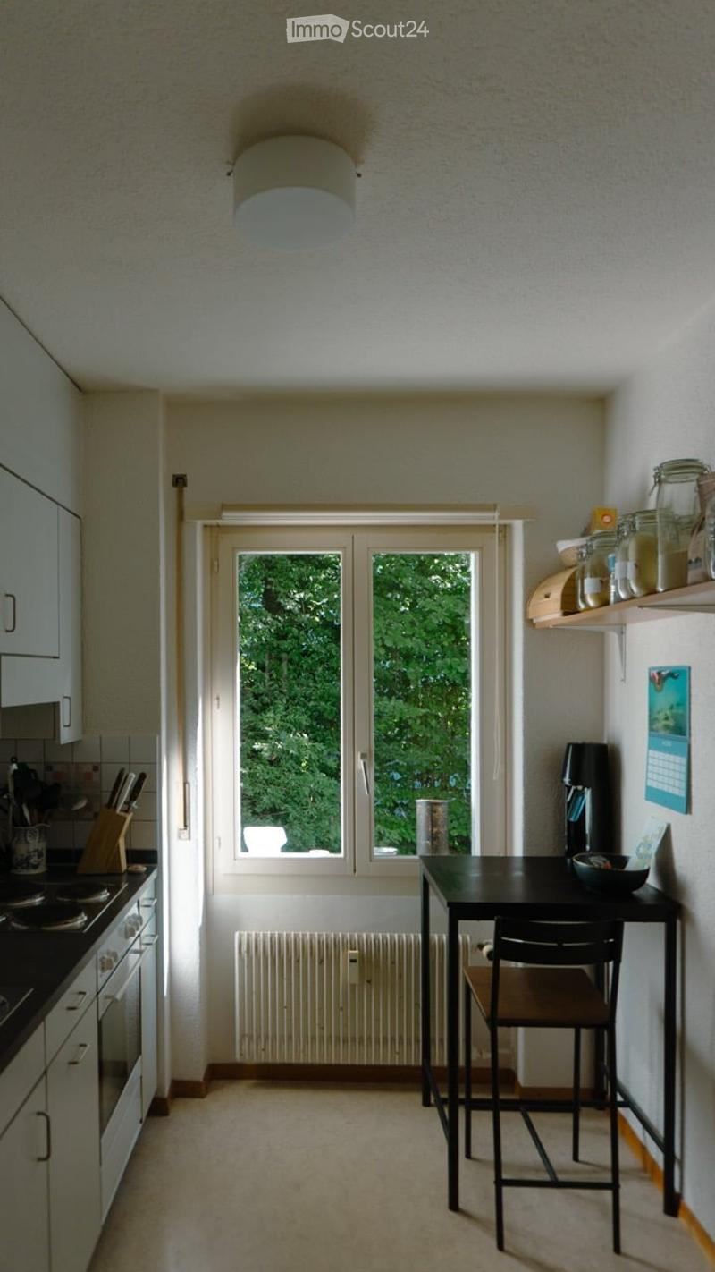 Bright kitchen with white cabinets, a black dining table and chair, a stove, and shelves with jars and a calendar.