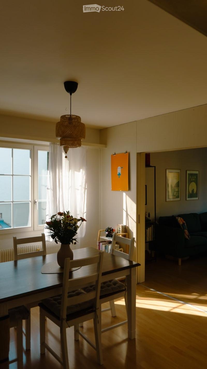 Bright dining area with wooden floor, large windows, dining table with chairs, vase with flowers, pendant lamp, and adjacent living space with sofa.
