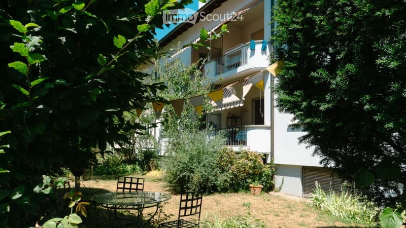 Garden area with a glass table and chairs, surrounded by trees and shrubs, with a building featuring balconies in the background.