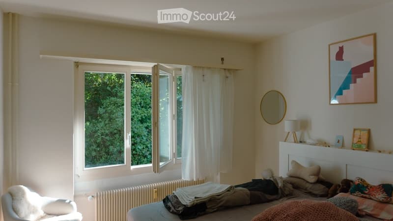 Cozy bedroom with a bed, a white chair, a round mirror, a small table lamp, and a colorful picture frame on the wall. Large windows with white curtains let in natural light.