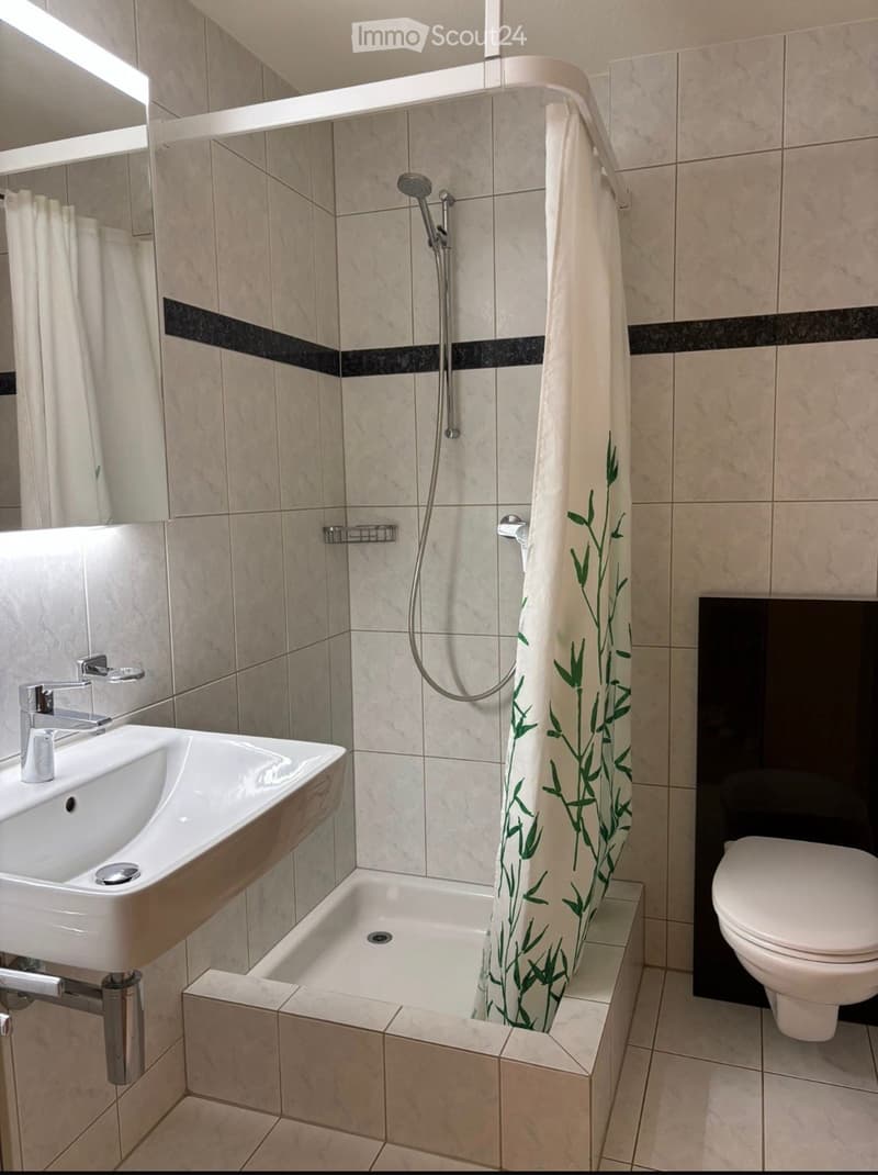 Modern bathroom with a white sink, chrome faucet, toilet, and a small shower with a bamboo-patterned curtain. The walls are tiled with beige tiles and black accents.