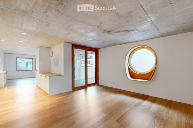 Empty room with wooden flooring, white walls, and large windows, including a round porthole window and a sliding glass door leading to another space.