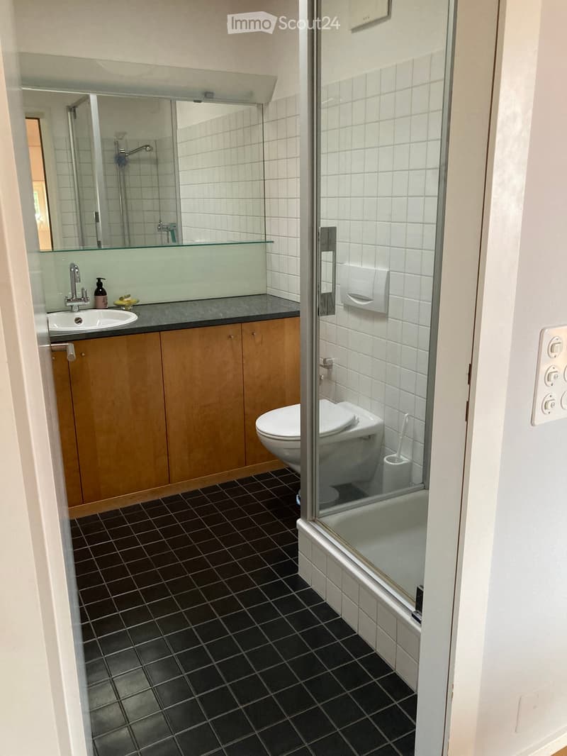 Modern bathroom with dark tiled floor, white tiled walls, wooden cabinetry, sink, toilet, and shower area with glass enclosure.