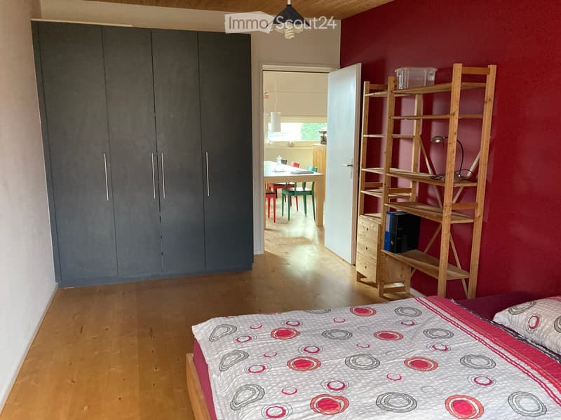 Bedroom with a bed, wardrobe, wooden shelf, and an open doorway leading to a dining area with a table and chairs.