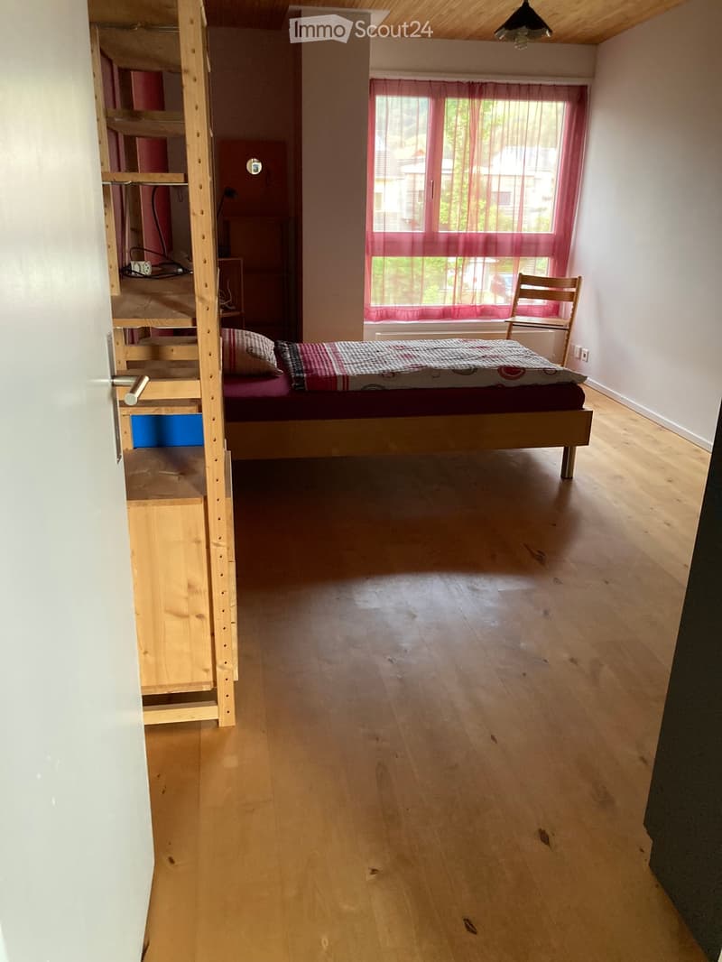 Cozy bedroom with wooden floor, single bed with patterned bedding, wooden chair near window with red curtains, and a bunk bed structure visible in the background.