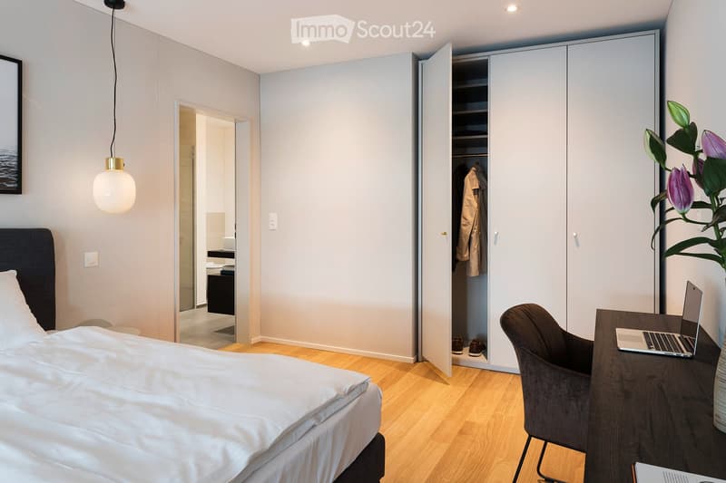 Modern bedroom with a large bed, a desk with a laptop, a chair, a wardrobe with clothes, and a plant in the corner.