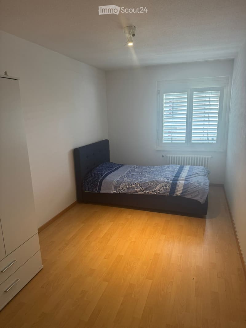Bedroom with wooden flooring, a single bed with dark headboard and patterned bedding, a wardrobe with silver handles, and a window with blinds.