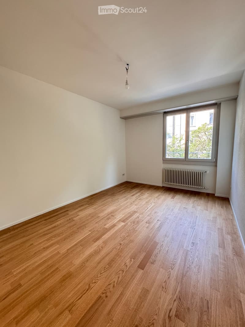 Empty room with wooden flooring, white walls, a single window with a radiator below, and a hanging light fixture.