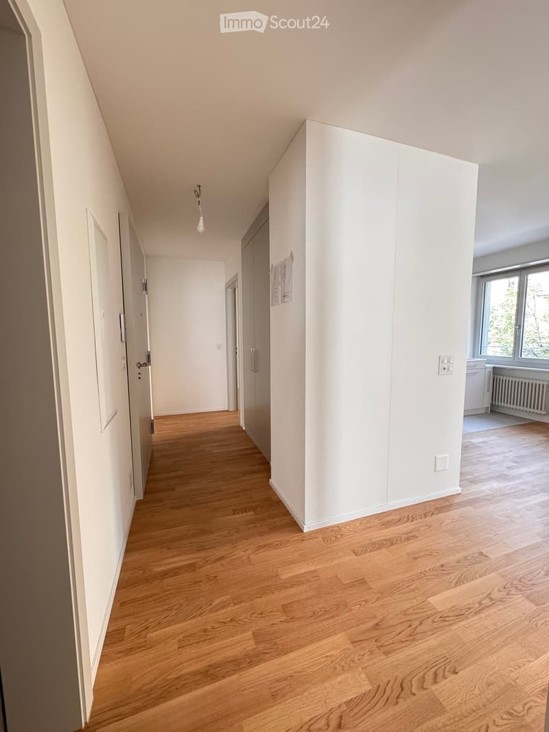 Empty hallway with wooden flooring, white walls, and several doors leading to other rooms. Natural light comes from a window at the end of the hallway.