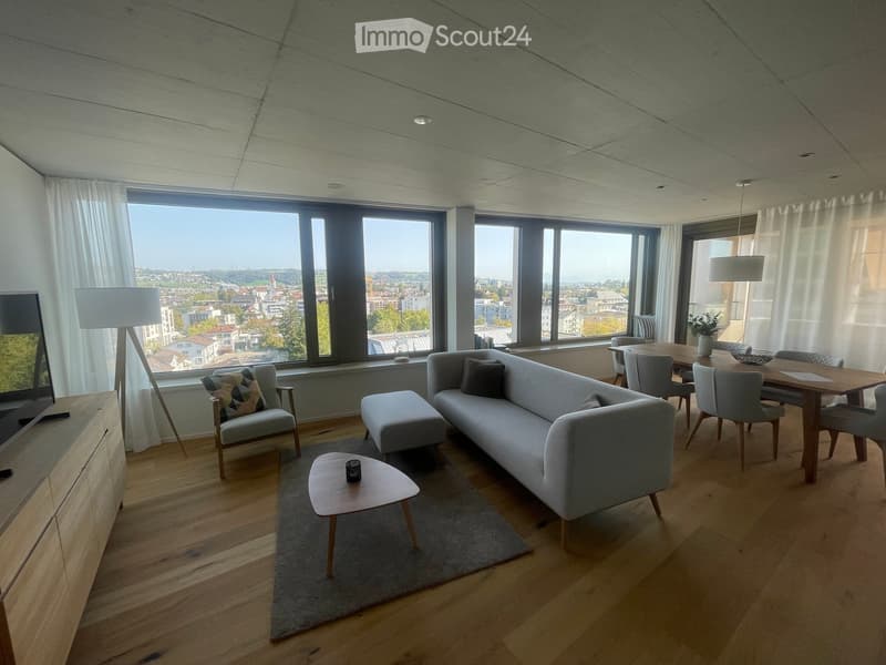 Modern living and dining area with large windows offering a city view, featuring a grey sofa, coffee table, ottoman, armchair, dining table with chairs, and floor lamps.