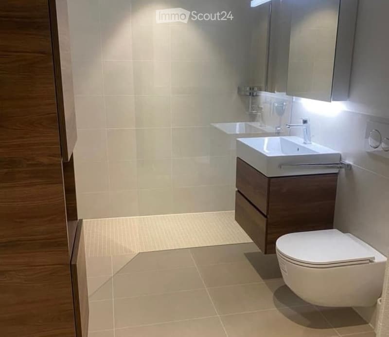 Modern bathroom with beige tiles, a white sink with a wooden cabinet, a toilet, and a mirror above the sink.