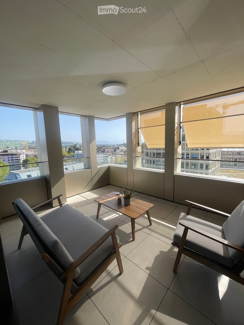 Modern balcony with tiled floor, large windows offering a city view, a sofa, a chair, a small table with plants, and roller blinds.