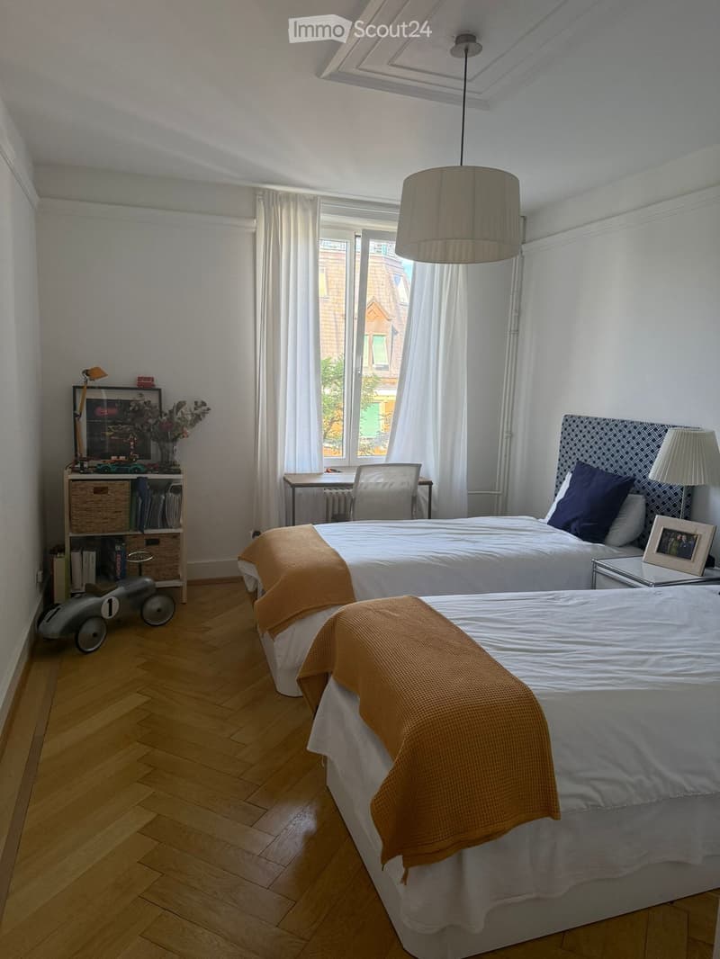 Bright bedroom with wooden flooring, two beds, nightstand, desk, chair, shelf, and decorative items.