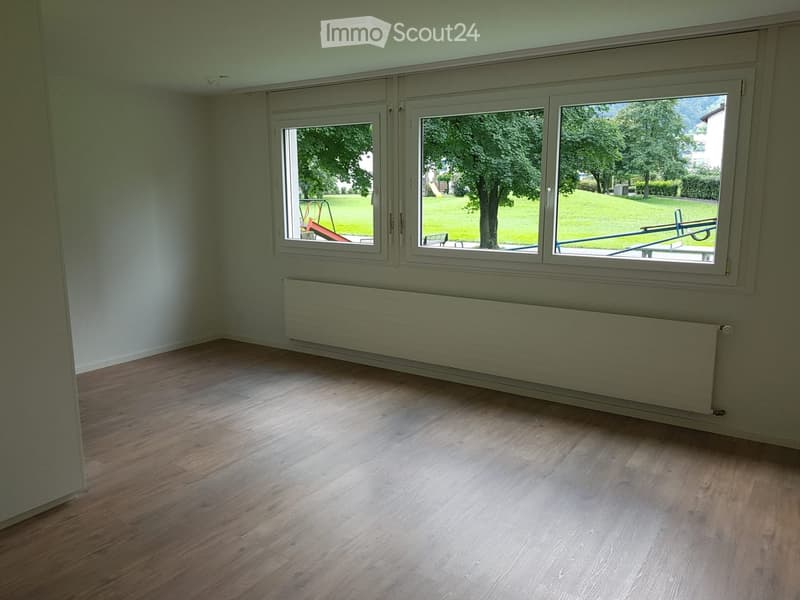 Empty room with wooden flooring, large windows, radiator, and view of a green outdoor area.