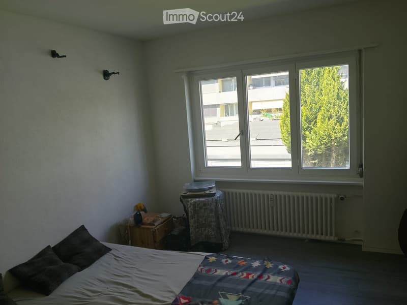 Bedroom with a bed, small table, radiator, and stool near the window.