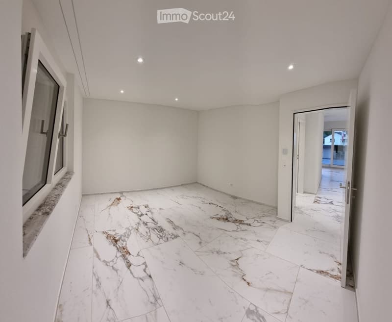 Empty room with white walls, marble flooring, and recessed lighting. Features a large window and an open doorway leading to another space.
