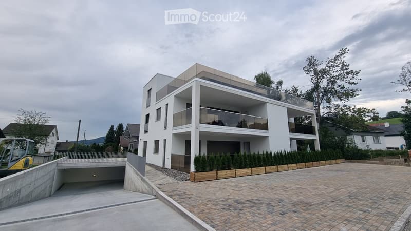 Modern two-story house with white facade, large windows, and balconies. Features a covered parking area and landscaped entrance.