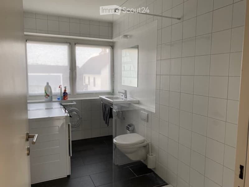 Modern bathroom with white tiled walls, a sink with a mirror, a toilet, a washing machine, and a cabinet.