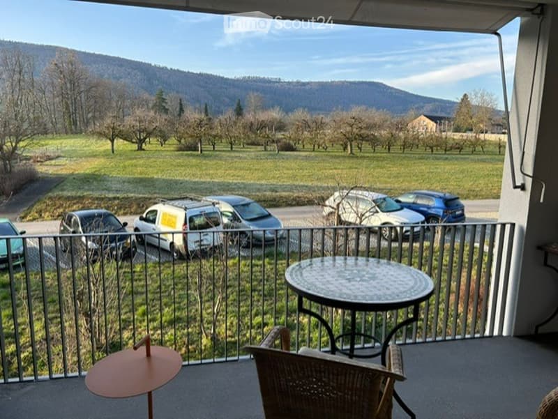 Balcony with a view of a green field, parking area, and distant mountains. Features a round table and a wicker chair.