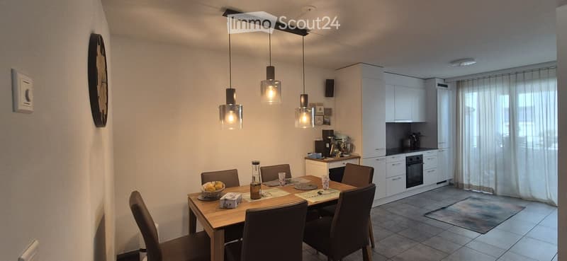 A modern dining and kitchen area with a wooden dining table, six chairs, a bowl of fruit, a bottle, and a cup on the table. The kitchen features white cabinets, an oven, a fridge, and various appliances.
