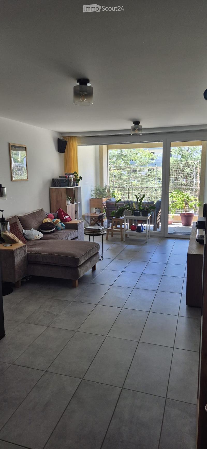 Modern living room with grey sofa, ottoman, small tables, and shelves. Large sliding glass doors open to a balcony with plants.