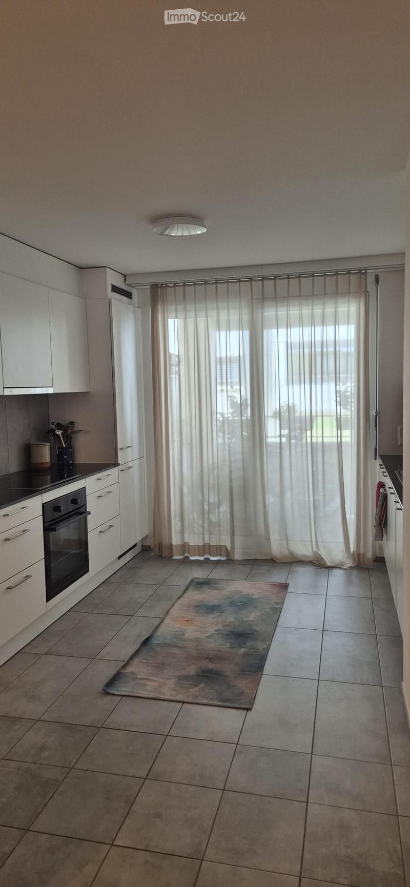Modern kitchen with white cabinets, an oven, refrigerator, and a large window with sheer curtains. A colorful abstract rug is placed on the tiled floor.