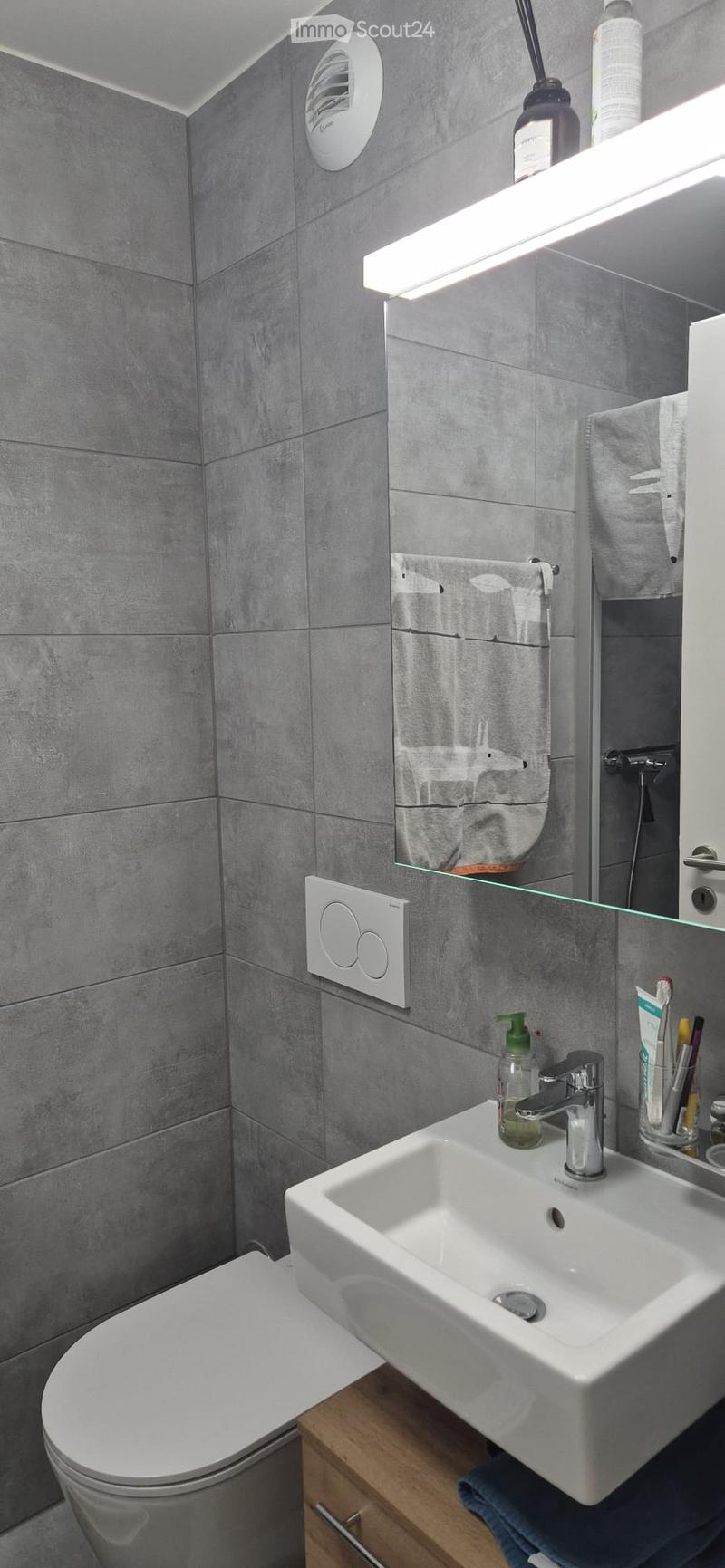 Modern bathroom with grey tiled walls, a white sink with a chrome faucet, a toilet, a large mirror with a built-in shelf holding a towel, and a toothbrush holder on the counter.