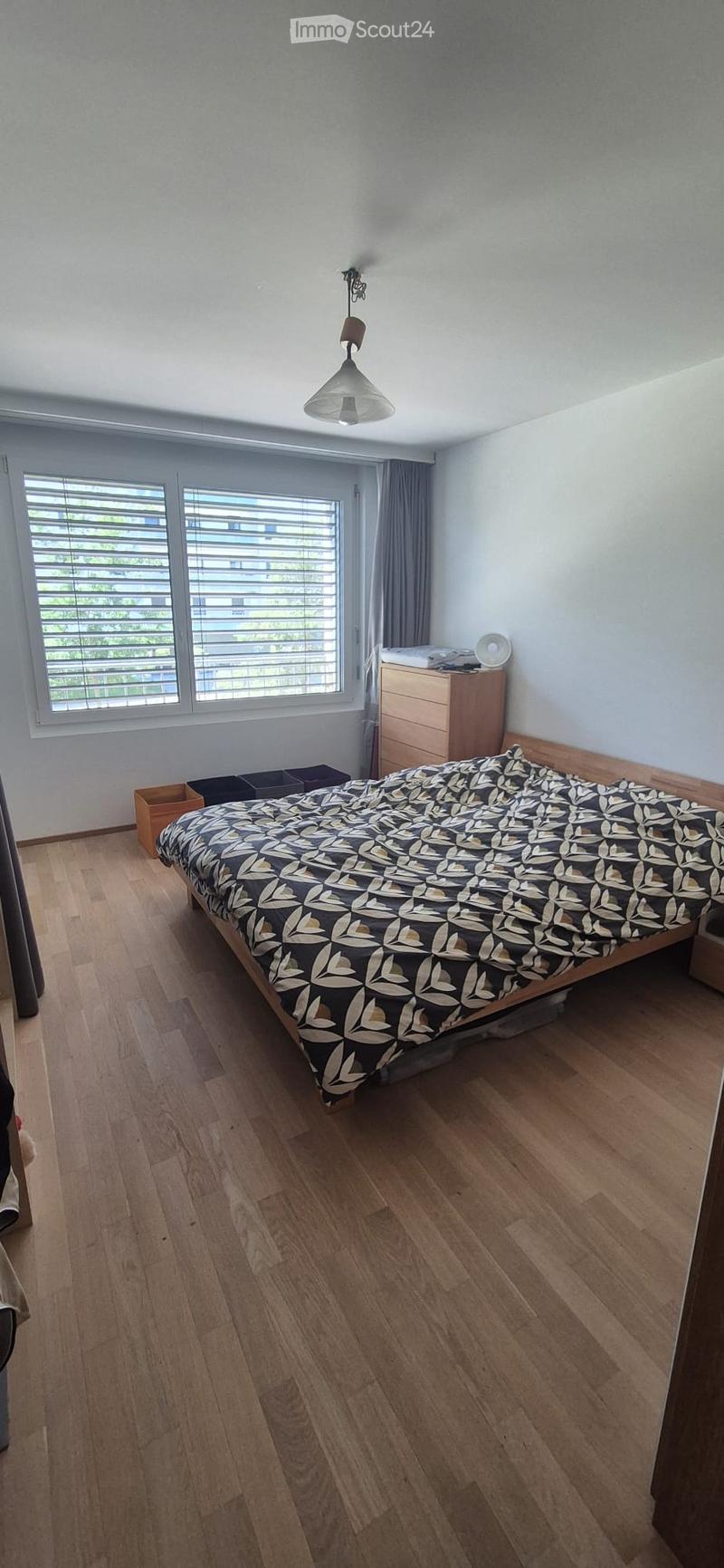 Bedroom with wooden flooring, large window with blinds, bed with patterned duvet, nightstand, and storage boxes.