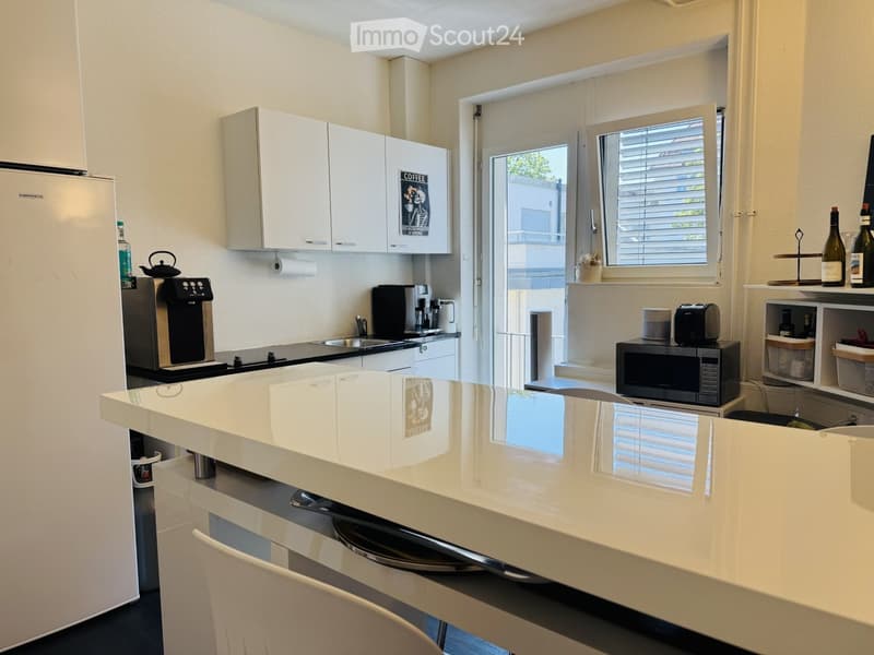 Modern kitchen with white cabinets, a central island, and various appliances including a coffee maker, microwave, and toaster. There is a window with blinds and a small shelf with bottles and a vase.