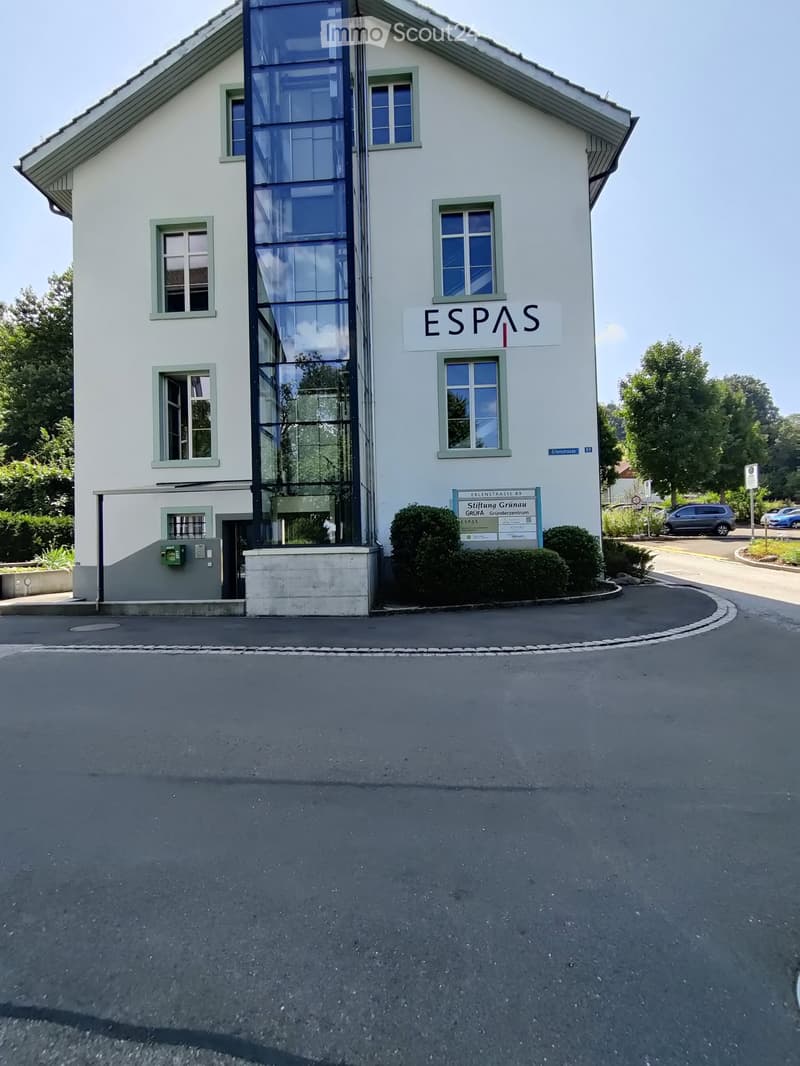 Two-story building with white facade, green-framed windows, and a modern glass elevator. Sign reads "ESPAS" and "Stiftung Grunau" with additional signage below.
