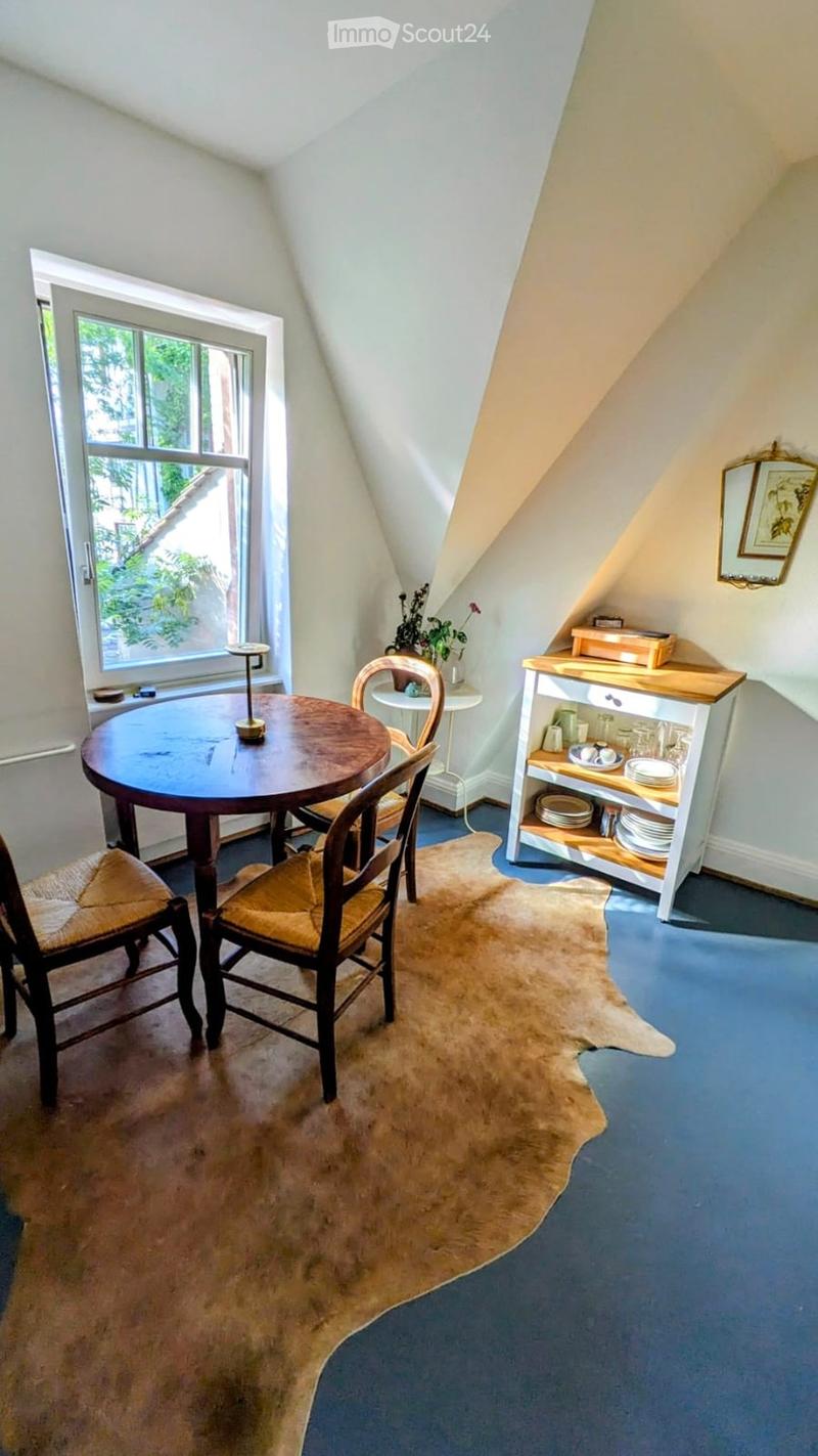 Cozy attic room with slanted ceilings, blue carpet, round wooden table, chairs, small cabinet, and decorative items.