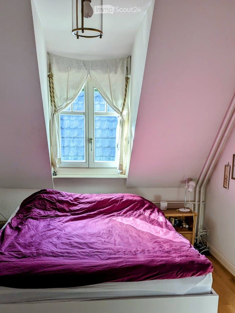 Cozy attic bedroom with slanted ceilings, a large window with white curtains, a neatly made bed with a purple duvet, a wooden nightstand with a lamp, and a small shelf holding books and decorative items.