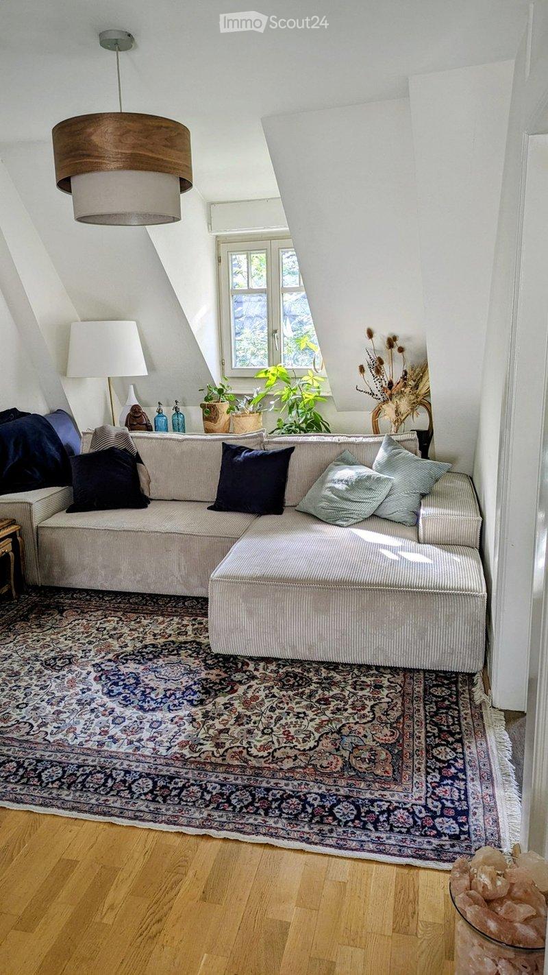 A modern living room with a large sectional sofa, ottoman, patterned rug, wooden floor, and large window with plants. The room features a hanging pendant light and decorative items on a side table.