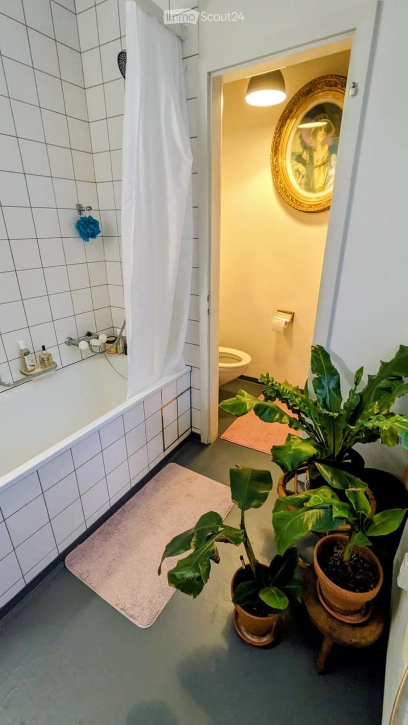 Bathroom with a bathtub, toilet, and potted plants near the entrance. The space features white tiled walls and a grey floor.