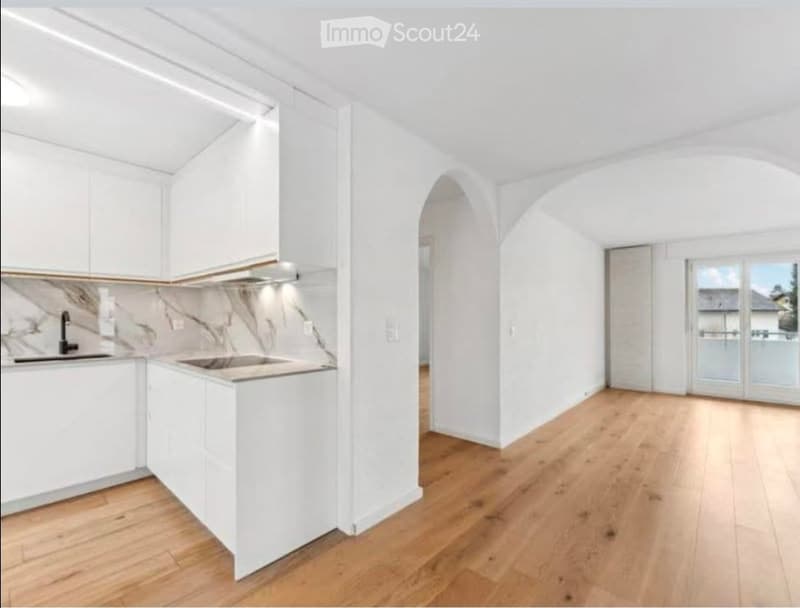 Modern kitchen with white cabinets, marble countertop, and wooden flooring, connected to a bright living space with large windows and sliding doors leading to a balcony.