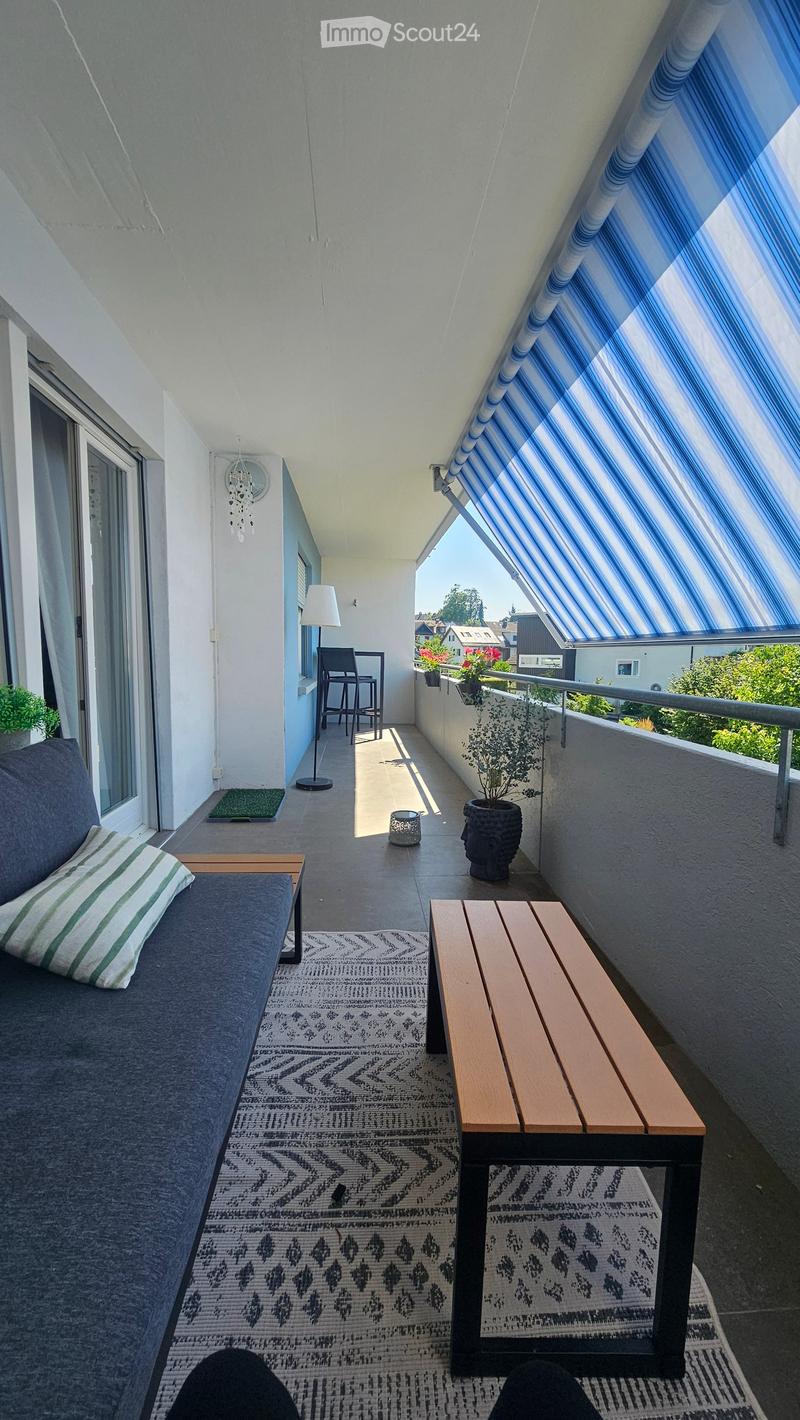 Balcony with a grey sofa, wooden bench, small table, chairs, floor lamp, rug, and potted plants.