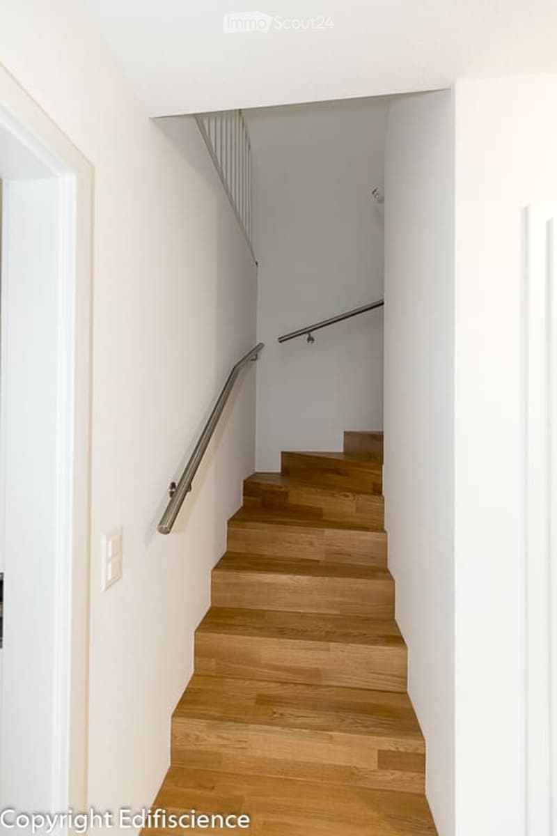 A modern, minimalist staircase with wooden steps and a metal handrail, set against white walls and a white ceiling.