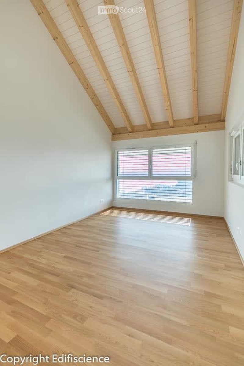 Empty room with wooden flooring and exposed wooden beams on the ceiling. Large window with blinds allows natural light to enter.